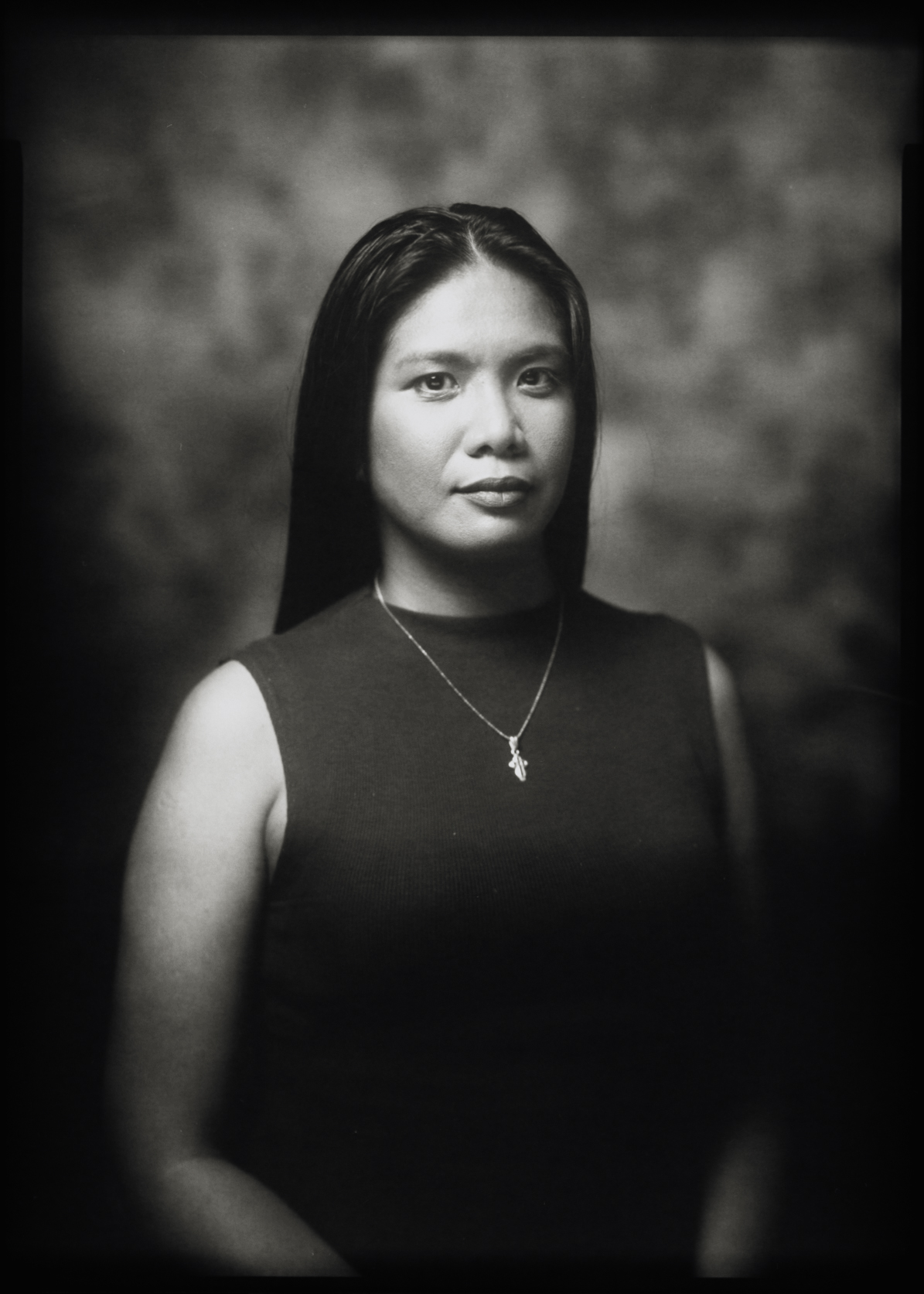 Black and white 5×7 large format studio portrait of a woman with long hair and a pendant necklace against a mottled backdrop